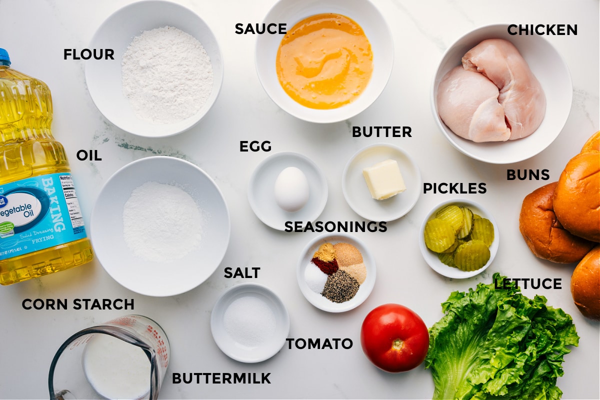 Ingredients In Crispy Chicken Sandwich All the ingredients prepped and ready for crispy chicken sandwich assembly, including oil, cornstarch, flour, sauce, egg, seasonings, salt, tomato, butter, pickles, lettuce, chicken, and buns.