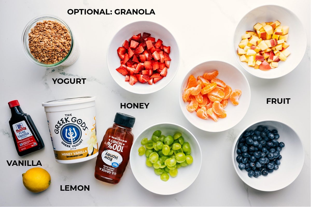 Ingredients In This Fruit Salad WIth Yogurt All ingredients prepped and ready for easy assembly including vanilla, lemon, honey, yogurt, strawberries, oranges, apples, blueberries, and grapes.