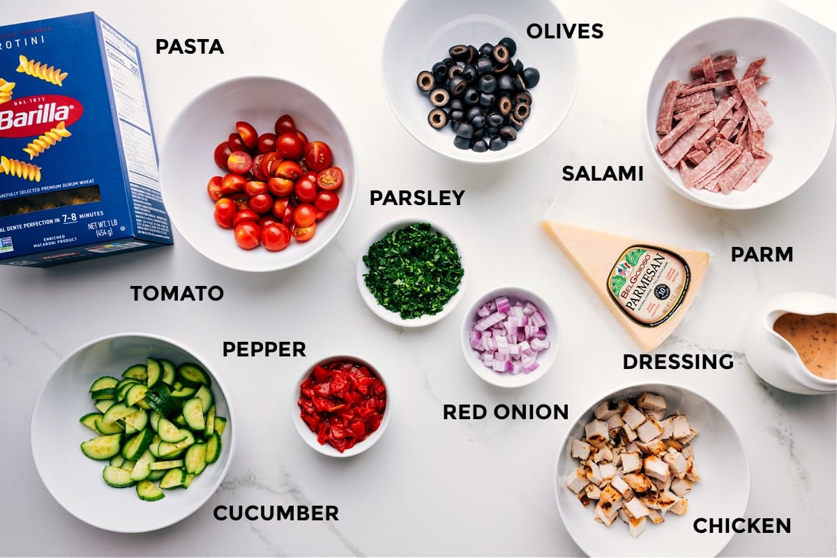 Ingredients In Italian Pasta Salad Recipe All the Italian Pasta Salad ingredients prepped for easy assembly, including pasta, olives, tomatoes, parsley, red onion, salami, Parmesan, dressing, chicken, cucumber, and roasted red pepper.