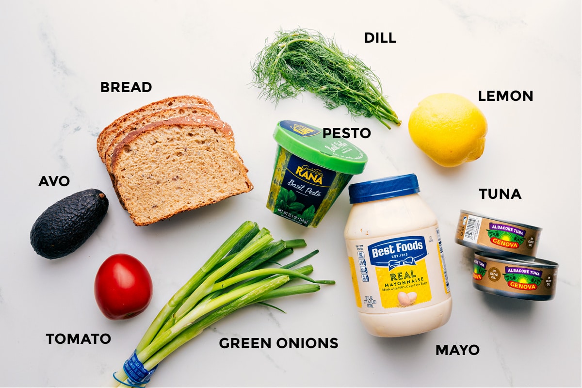 Ingredients In Tunacado All the ingredients prepped for easy assembly, including avocado, tomato, bread, green onions, pesto, dill, lemon, tuna, and mayo.