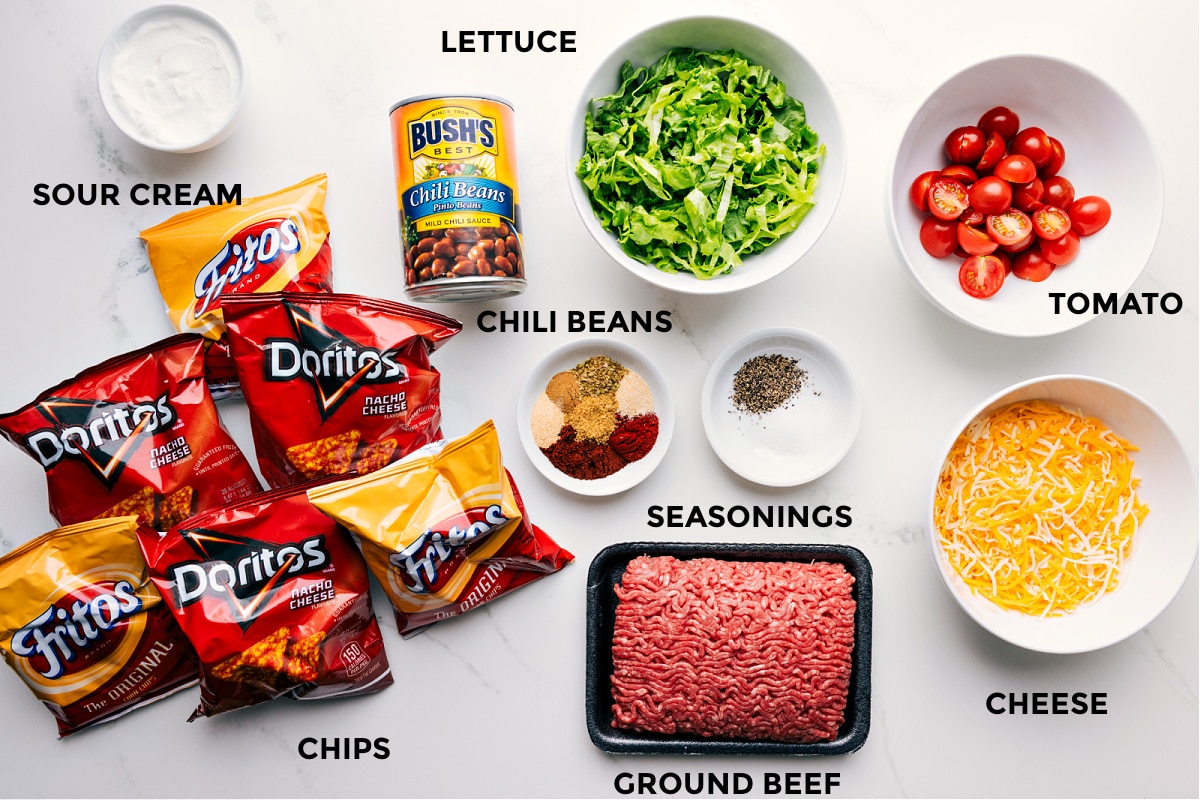 Ingredients In Walking Taco Recipe Ingredients in this recipe prepped for easy assembly, including sour cream, chili beans, lettuce, tomato, cheese, seasonings, ground beef, and chips.