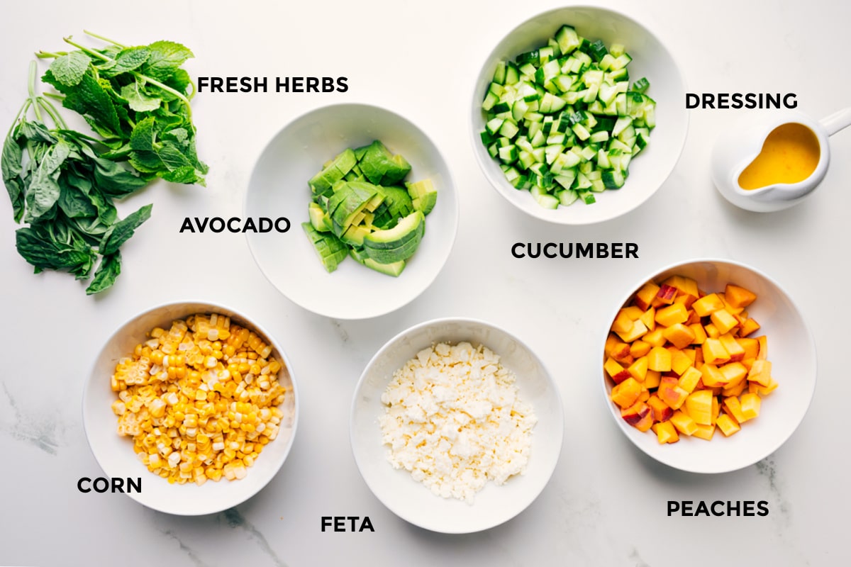 Ingredients In Peach Feta Salad Ingredients in this recipe prepped out for easy assembly including the fresh herbs, avocado, corn, feta, cucumber, peaches, and dressing.