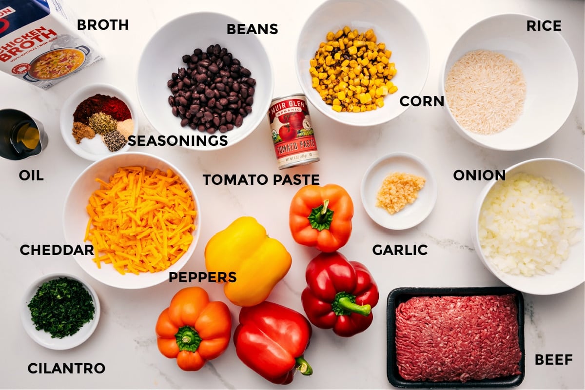 Ingredients In Mexican Stuffed Peppers Stuffed pepper ingredients prepped for easy assembly, including broth, oil, spices, cheddar cheese, cilantro, peppers, beans, tomato paste, garlic, ground beef, rice, corn, and onions.