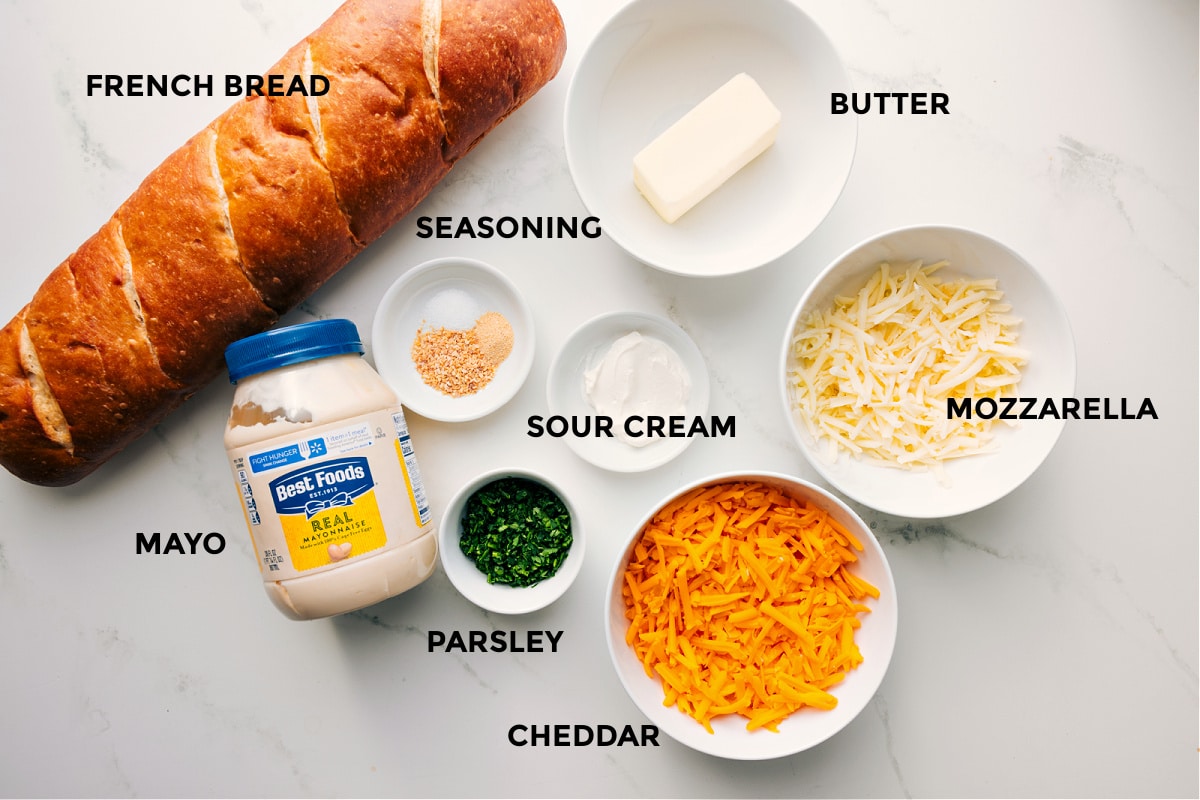 Ingredients In Cheesy Bread Recipe All the ingredients prepped and ready to assemble, including French bread, mayo, seasonings, parsley, sour cream, butter, mozzarella, and cheddar.