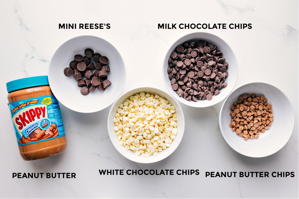 Ingredients In Peanut Butter Bark Ingredients prepped in small bowls for easy assembly including peanut butter, mini Reeseโs cups, white chocolate chips, milk chocolate chips, and peanut butter chips.