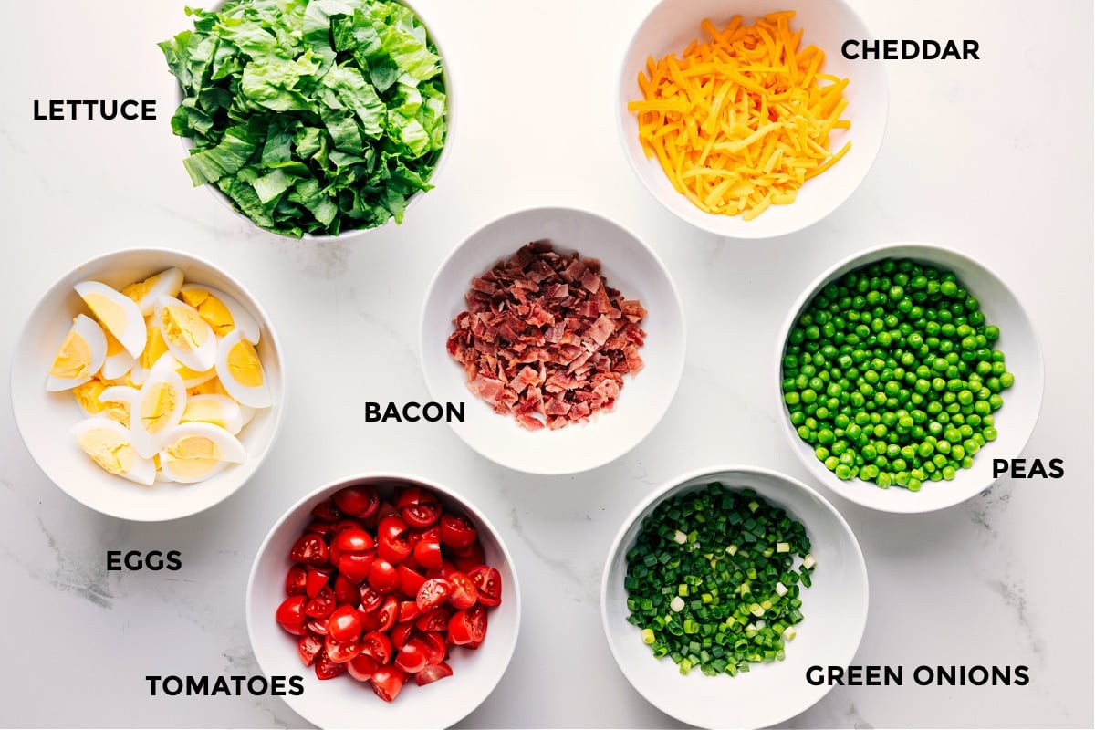 Ingredients In This 7 Layer Salad All the ingredients in this recipe prepped out for easy assembly including the lettuce, eggs, tomatoes, bacon, green onions, peas, and cheddar.