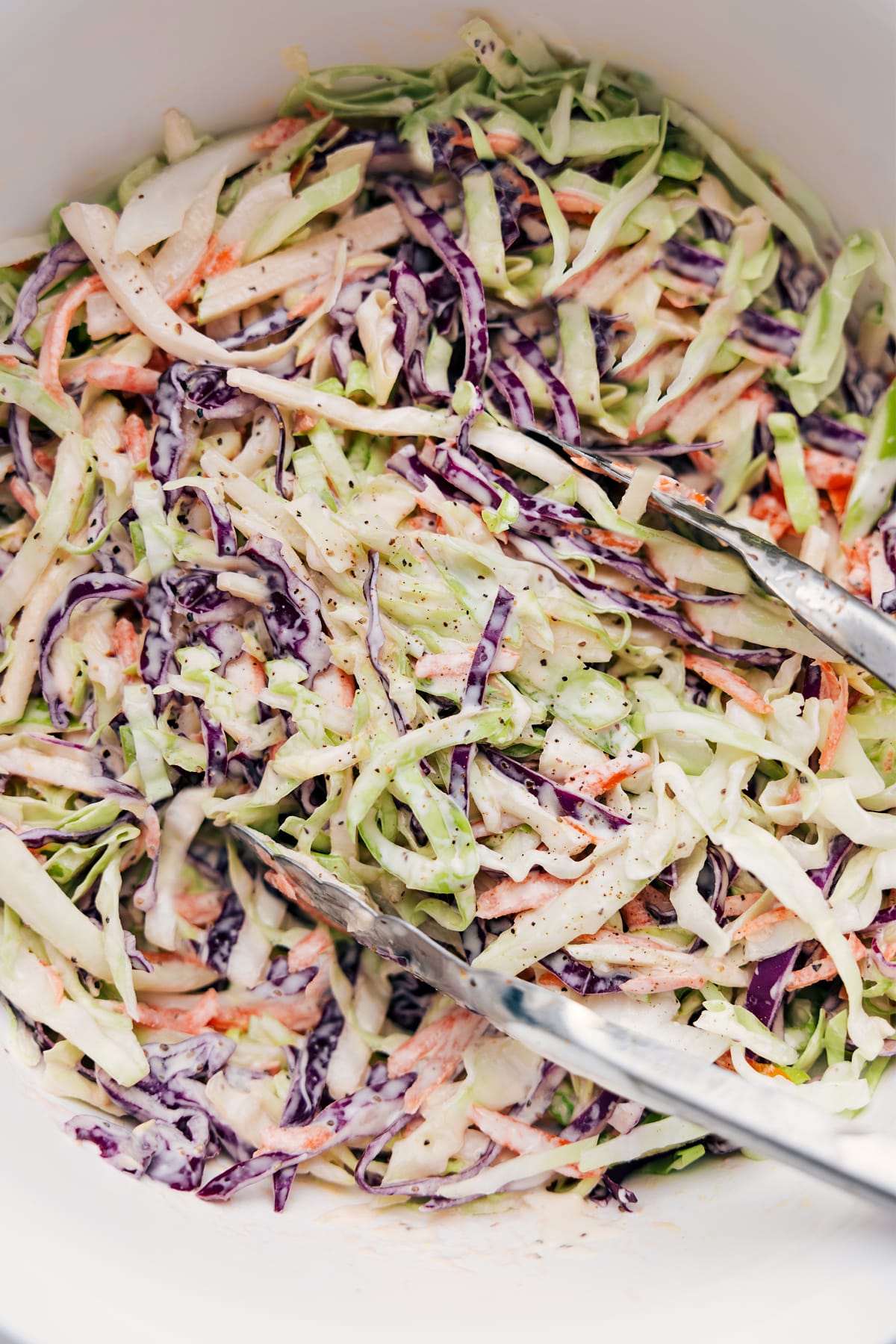 Easy Coleslaw Recipe being tossed in a creamy dressing, with tongs lifting a large scoop.