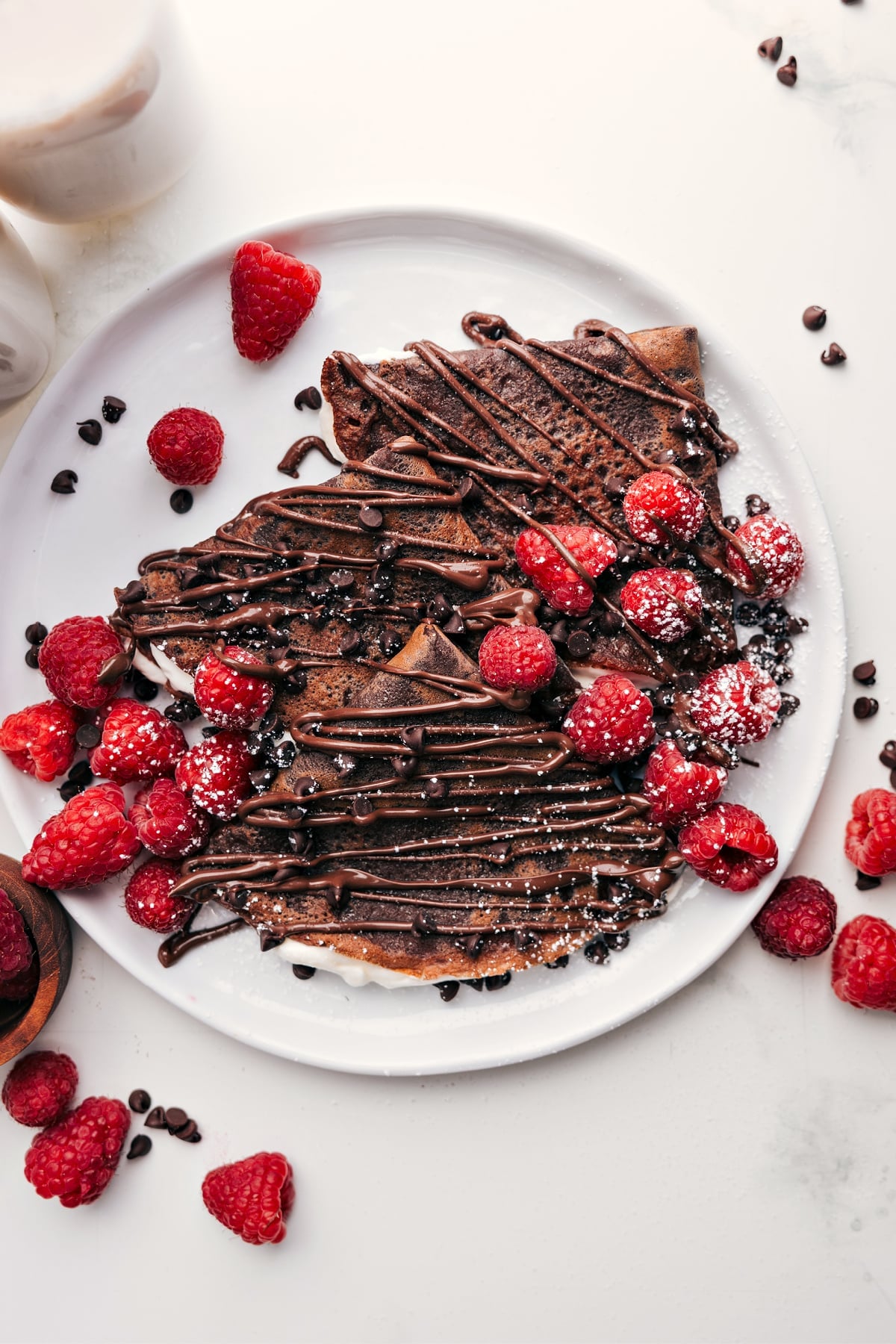 Chocolate Crepes topped with chocolate drizzle, mini chocolate chips, fresh berries, and a dusting of powdered sugar on a plate.