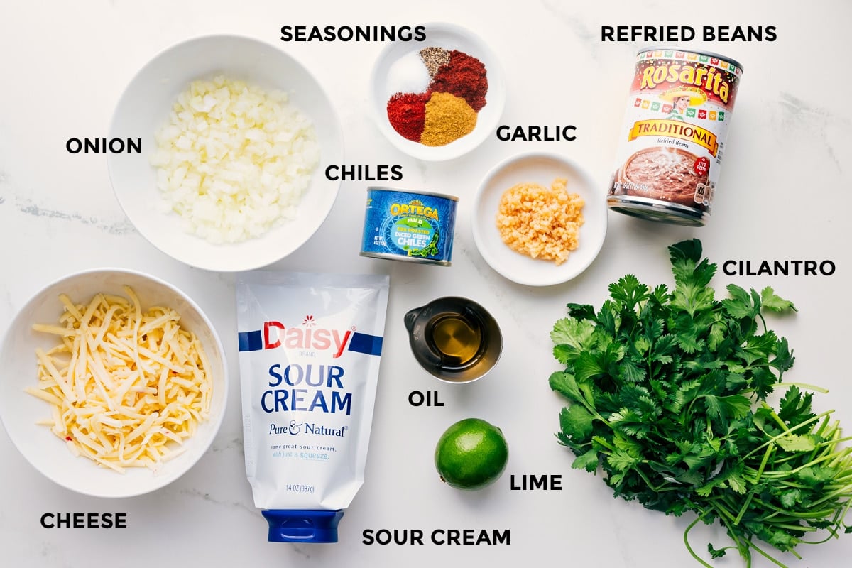 All the ingredients in this recipe prepped out for easy assembly including the cheese, onion, sour cream, lime, oil, chiles, seasoning, garlic, refried beans, and cilantro.
