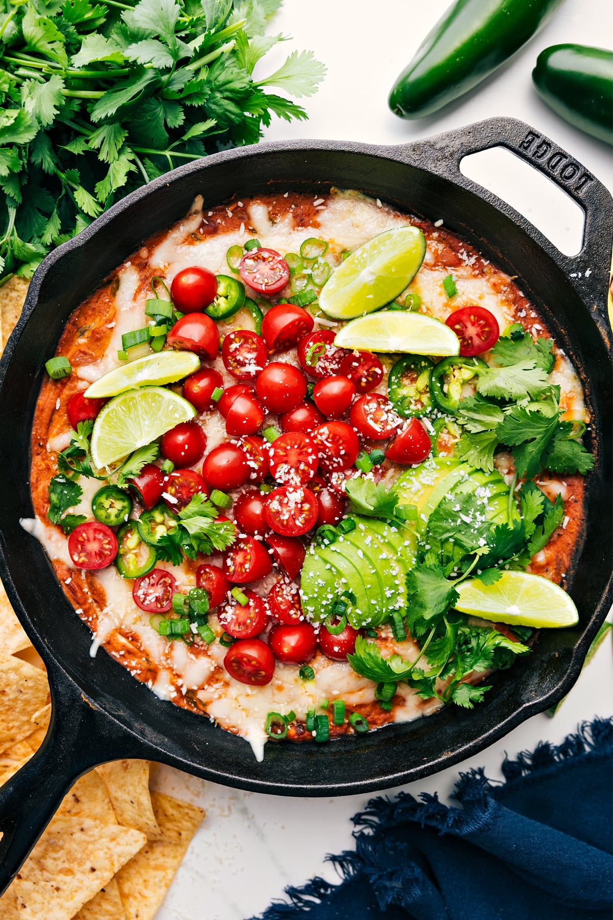 Refried Bean Dip in a skillet topped with fresh tomatoes, avocado, lime wedges, green onions, and cilantro.